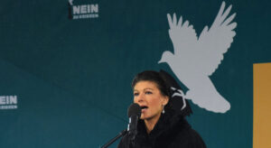 Sahra Wagenknecht mit Mikrofon auf einer Kundgebung. Im Hintergrund zu sehen eine Friedenstaube und der Slogen "Nein zu Kriegen".