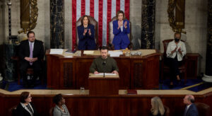 People stand in an old hall on wooden stands and applaud a man who is giving a speech. An american flag in the background on the wall.