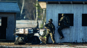 Image shows three soldiers, one of them climing into the window of a small wooden building, the other two holdling guns, next to a small vehicle with a flat top that holds several devices.