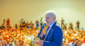 President Lula, wearing a blue suit and holding a wireless microphone, speaking in front of a crowd that is drenched in warm light.