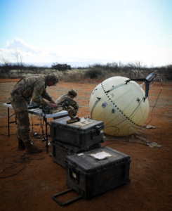 Zwei US-Soldaten in Uniform basteln an einer Art Ballon-Drohne. Sie befinden sich im freien Gelände in der Halbwüste