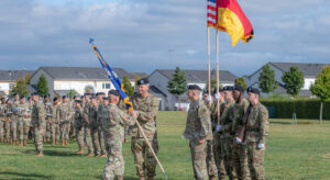 Ein Soldat überreicht lächelnd einem anderen Soldaten eine blaue, zusammengewickelte Fahne. Rechts von ihnen stehen sechs Soldat*innen, von denen zwei jeweils die deutsche und die US-amerikaische Fahne tragen. Sie befinden sich auf einer grünen Wiese, im Hintergrund sind links weitere Soldat*innen zu sehen. Dahinter befinden sich weiße Reihenhäuser.