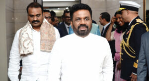A man formally dressed in white walks towards the camera, followed by other formally dressed people. Others stand on the side including people dressed in military uniform