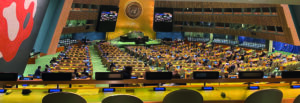 The large conference room of the UN General Assembly with the UN logo on the wall above the podium.