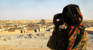 Person with black and red patterned headscarf seen from behind overlooking a settlement through binoculars.