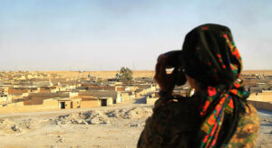 Person with black and red patterned headscarf seen from behind overlooking a settlement through binoculars.