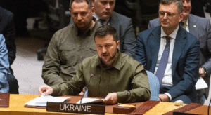 Volodymyr Zelenskyy sitting at a conference desk and speaking into a microphone. In front of him on the desk is a nameplate with "UKRAINE" written on it.