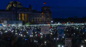 Das Bild zeigt eine große Menschengruppe im dunklen vor dem Bundestag. Sie halten Lichter in die Höhe und Schilder, die Protest gegen die AfD ausdrücken.