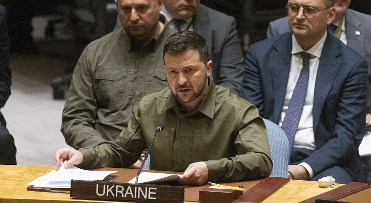 Volodymyr Zelenskyy sitting at a conference desk and speaking into a microphone. In front of him on the desk is a nameplate with "UKRAINE" written on it.