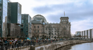 Eine große Gruppe Menschen mit Plakaten, die die Ukraine-Flagge zeigen, laufen links nebem einem Fluss entlang. Im Hintergrund ist das Gebäude des Deutschen Bundestags mit der Glaskuppen zu sehen.