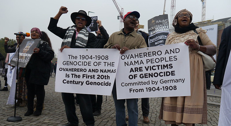 Protesters holding up signs, the visible signs read: The 1904-1908 genocide of the Ovaherero and Namas is the first 20th century genocide, and: Ovaherero and Nama peoples are victims of genocide committed by Germany from 1904-1908.