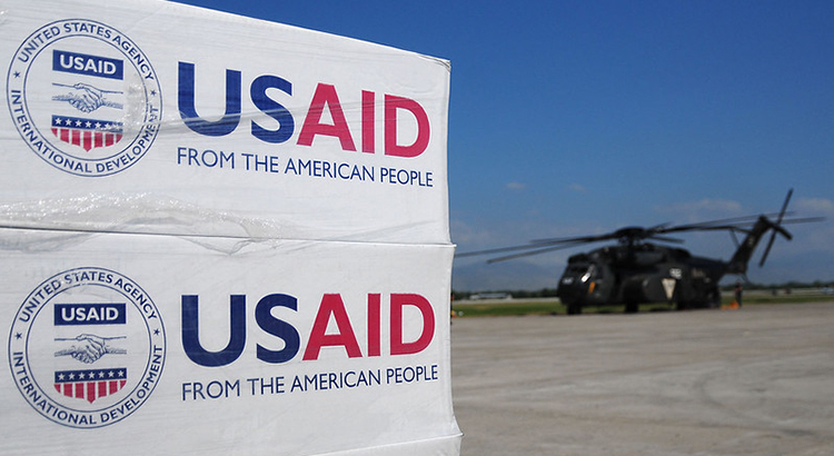 Stack of boxes with the USAID logo and writing "USAID. From the American people." in the foreground. In the background there is an airfield with a helicopter.