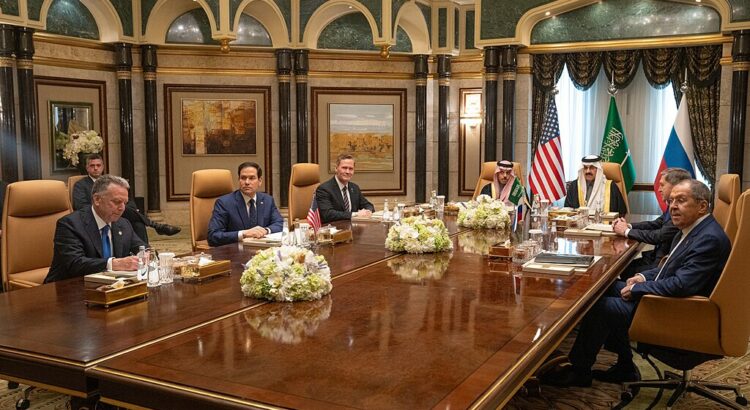 Photo einer Gruppe von Männern, die in einem prunkvollen Saal an einem Tisch sitzt. Dahinter sieht man die Flaggen von USA, Saudi-Arabien und Russland