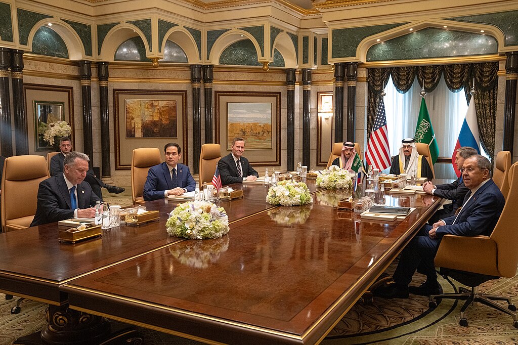 Photo einer Gruppe von Männern, die in einem prunkvollen Saal an einem Tisch sitzt. Dahinter sieht man die Flaggen von USA, Saudi-Arabien und Russland