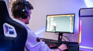 Ein Mann mit Kopfhörern sitzt vor einem Gamer-Bildschirm auf dem man diffus Chats erkennen kann