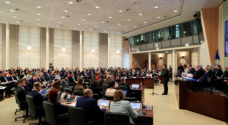 Room with semi-circular tables at which formally dressed people sit. A man in a suit is standing at the lectern, with a number of other people sitting behind him. The flag of the OPCW hangs on the wall.