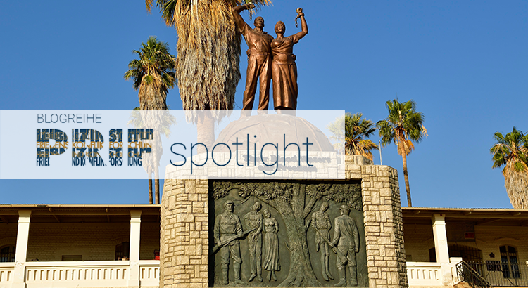 The picture shows the Genocide Memorial in Windhoek, Namibia which commemorates the extermination order against Ovaherero and Nama following their rising aganist German rule in Namibia. The Genocide memorial is made of stone with a plaque showing people being hanged and german officers with guns standing in front of them. Atop the memorial two bronze figures raise their fists. On the stone it reads “Their Blood Waters our Freedom”