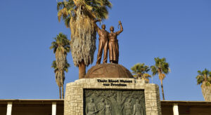 The picture shows the Genocide Memorial in Windhoek, Namibia which commemorates the extermination order against Ovaherero and Nama following their rising aganist German rule in Namibia. The Genocide memorial is made of stone with a plaque showing people being hanged and german officers with guns standing in front of them. Atop the memorial two bronze figures raise their fists. On the stone it reads “Their Blood Waters our Freedom”