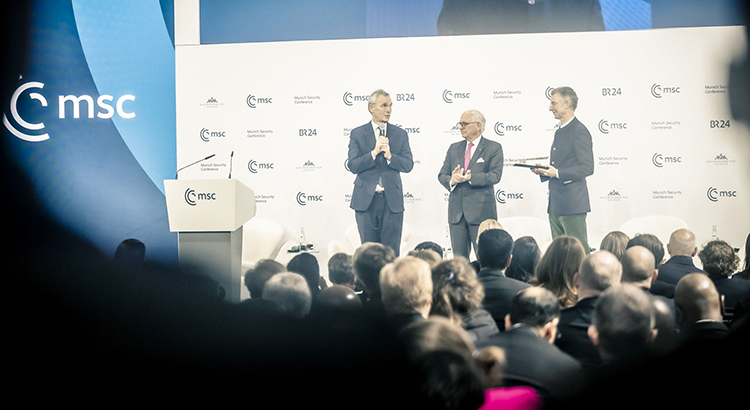 Photo of three men in suits on a stage in front of a crowd of people. To their left is a blue wall with the msc logo.