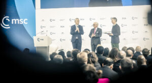 Photo of three men in suits on a stage in front of a crowd of people. To their left is a blue wall with the msc logo.