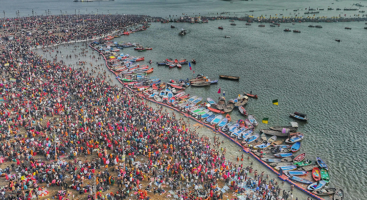 Photo of a beach filled with crowds of people in colorful clothes. Close to the shore there is a line of colorful boats.