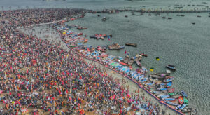 Photo of a beach filled with crowds of people in colorful clothes. Close to the shore there is a line of colorful boats.
