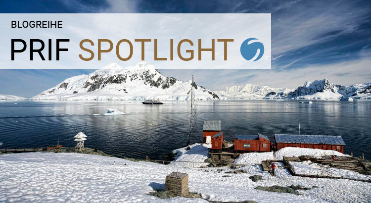 Antarctic coastline with snow mountain range visible in the distance. Three small red buildings are visible in the front right lower corner.