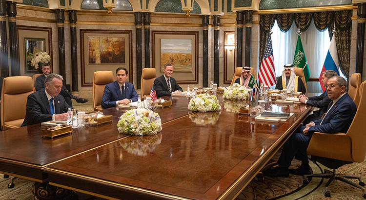 Photo of a group of men sitting at a table in a magnificent hall. Behind them you can see the flags of the USA, Saudi Arabia and Russia