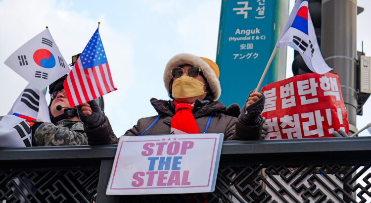 Frau mit Mütze, Sonnenbrille und Atemschutzmaske steht an einem Geländer, vor ihr hängt ein Schild mit Aufschrift "Stop the Steal". Sie hält in enier Hand eine südkoreanische und in der anderen Hand eine amerikanische Flagge. Daneben sind weitere Flaggen und Schilder mit koreanischen Schriftzeichen zu sehen.