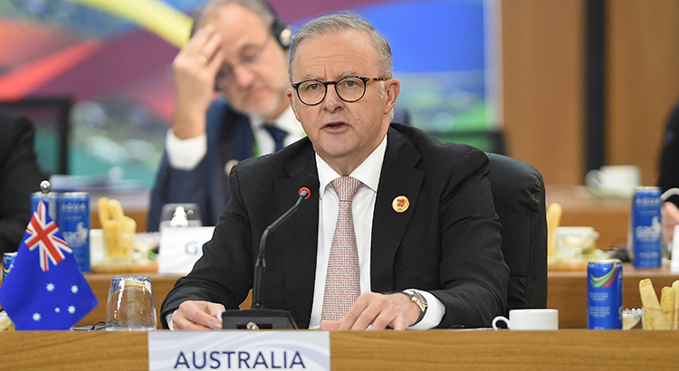 A man in a suit sits at a table with a microfone in front of him. next to him stands a tiny australian flag.