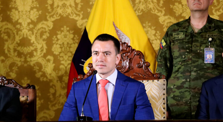 Daniel Noboa sitzt in einem prunkvoll aussehenden Raum auf einem Stuhl, hinter ihm sind eine Flagge von Ecuador und ein Mann in Militäruniform zu sehen.