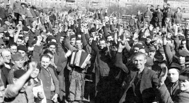 Schwarz-Weiß Foto. Eine große Gruppe Männer steht im Kreis. Sie lachen und reißen die Arme und Fäuste in den Himmel, ein Man trägt ein Akkordeon.