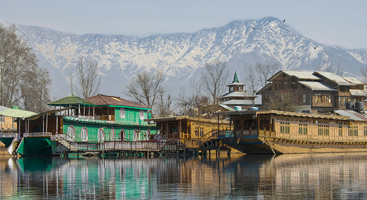 Photo of a green and a brown wooden house by a lake on which houseboats are floating. Snow-capped mountains rise up in the background.
