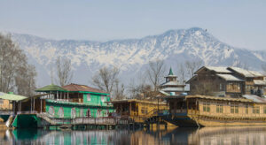 Photo of a green and a brown wooden house by a lake on which houseboats are floating. Snow-capped mountains rise up in the background.