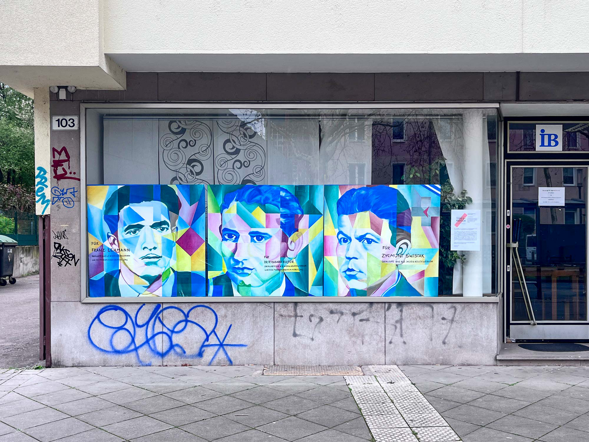 Ein Schaufenster. Bunte Porträts mit geometrischen Formen von drei Männern, die das KZ Katzbach überlebt haben.