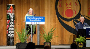 A woman stands on a stage with indigenous insigna behind her