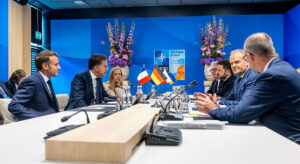 Auf beiden Seiten eines Konferenztisches sitzen fünf Staats- und Regierungschefs europäischer Staaten, der NATO Generalsekretär und der ukrainische Präsident. Zwischen Ihnen stehen Flaggen der jeweiligen Länder, im Hintergrund hängt das Poster des NATO Gipfels 2025