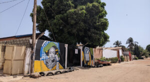 across the unpaved street there is a mural on a wall and a large tree.