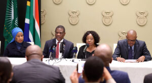Four people sit at a table facing the press and talk into microfones. In the background is a South African Flag.