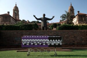 A purple-blue casket on a meadow in front of a statue of Nelson Mandela in Pretoria, South Africa. In the picture, it appears as if Mandela spreads his hands over the casket.
