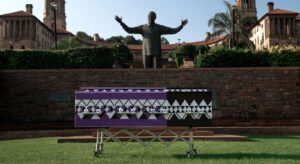 Black and purple patterned coffin from the campaign on a meadow in Cape Town, South Africa, with a statue of a man with his arms outstretched behind it. In the photo, it looks as if he is spreading his arms over the coffin.