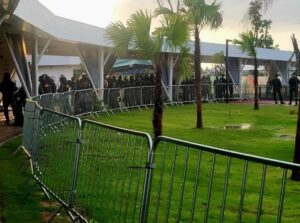 Bild zeigt eine Grüne Wiese mit Palmen darauf, die mit einem Zaun von einem weiß überdachten Gang abgegrenzt ist. Im Gang steht eine Gruppe schwarz gekleideter Menschen am Zaun.