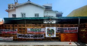 Bild des Hauses 'COP DO POVO' ("COP des Volkes"): Zu erkennen ist das Obergeschoss eines weißen Gebäudes, das sich hinter einem überdachten Holzzaun befindet. Am Zaun hängen Protestbanner zivilgesellschaftlicher Gruppen mit portugiesischer Aufschrift.