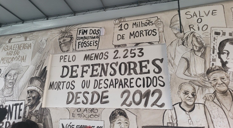 Wandmalerei im Haus 'COP DO POVO'. Gemalt wurden protestierende Menschen, im Mittelpunkt des Bilds ein Banner mit Aufschrift 'Pelo Menos 2.253 Defensores Mortos ou Despaparecidos desde 2012'.
