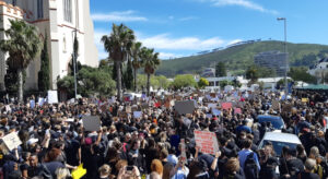 A crowd on a street in Cape Town in front of a mountain panorama. Most of the demonstrators are female and are holding up protest signs. One sign in the foreground reads in red letters: 'There is no place on earth more dangerous to women than South Africa.'