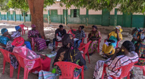Group of people sitting in a circle on plastic chairs outside underneath a tree.