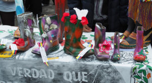Boots decorated with various motifs such as flowers and butterflies are standing on a table outdoors, behind which you can see people wearing jackets.