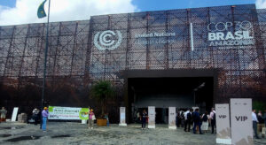 Der Zugangsbereich zur Blue Zone auf der COP30: Abgebildet ist ein Gebäude mit Aufschrift 'COP 30 Brazil - Amazonia'. Das Gebäude hat ein großes Tor, vor dem Menschen in zwei Schlangen stehen. Am Rand stehen Aufsteller mit der Aufschrift 'VIP'. Links vom Tor ist außerdem eine kleine Protestgruppe zu sehen, deren Banner für eine klimafreundliche, pflanzliche Ernährung wirbt.