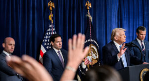 Donald Trump stands at a lectern next to the US flag. Government officials can be seen next to him, with a journalist's raised hand in the foreground.