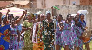 Group of colorfully dressed women walking. One woman is holding a drum, another a silver walking stick with a figure on top.